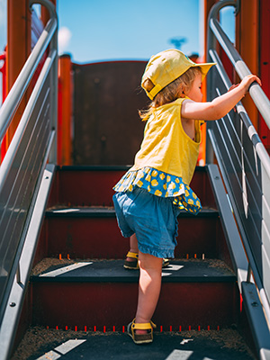 A llittle girl, toddler age, is climbing up small steps to get to a playground unit platform.